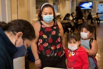 Community schools A woman and two young children wearing masks stand behind a table, looking down at something